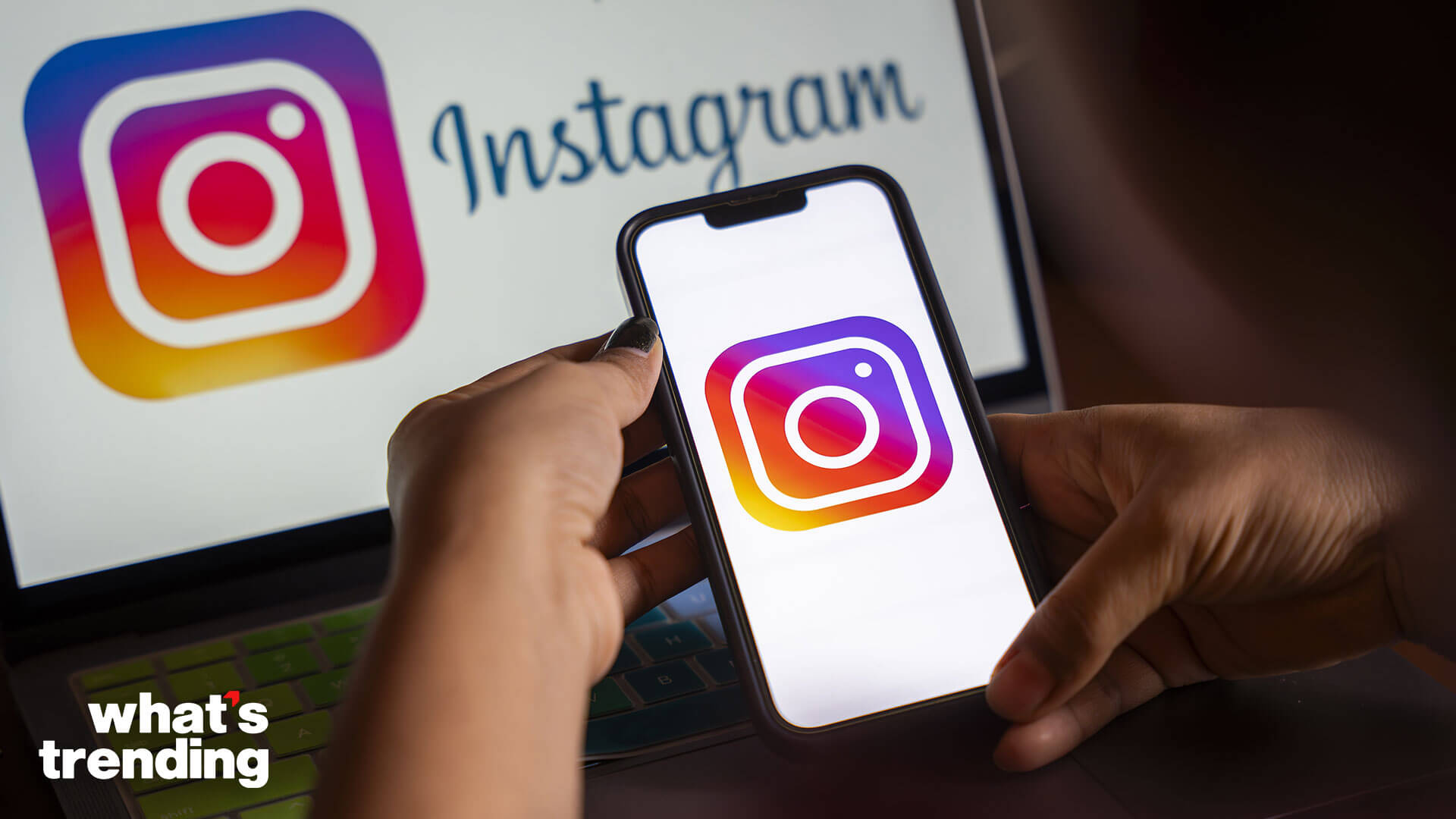 Hands holding a smartphone displaying the Instagram logo with a laptop showing the Instagram logo and text in the background